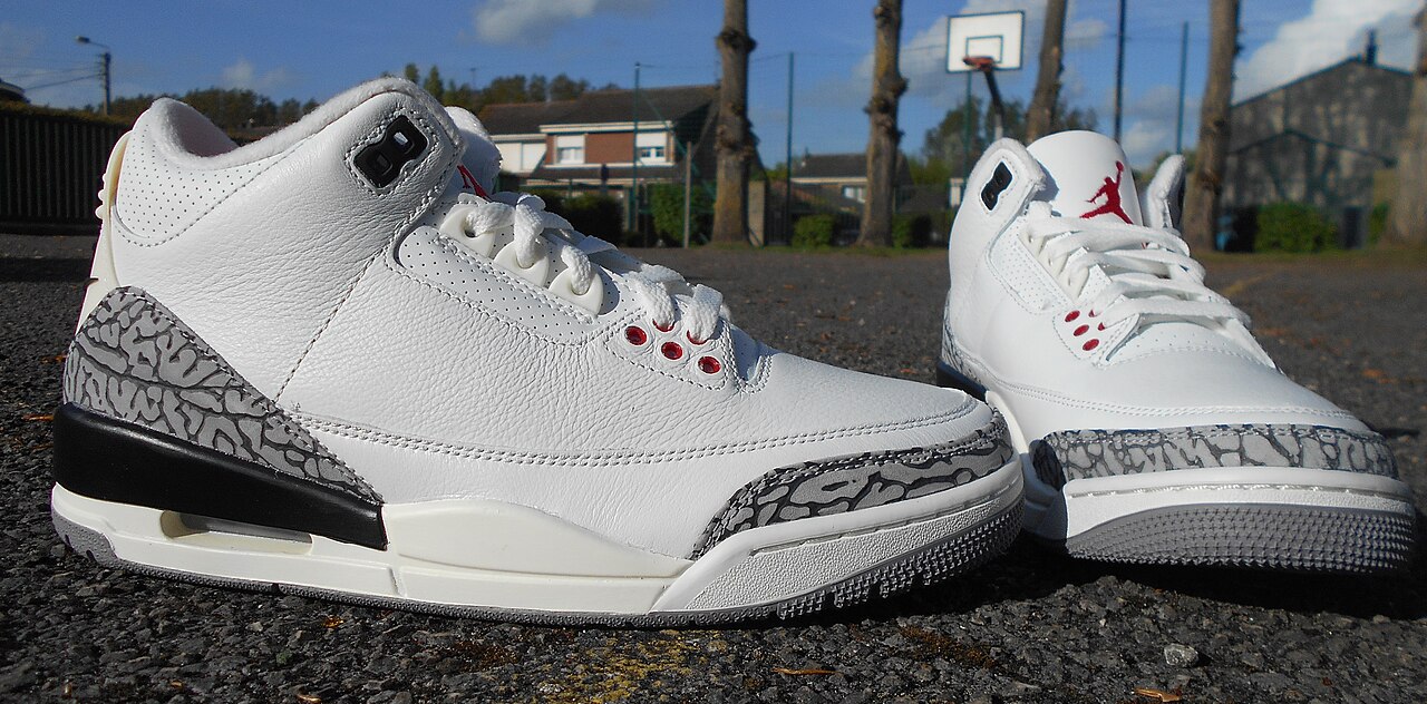 A pair of white Air Jordan 3 White Cement Reimagined sneakers with gray elephant-print panels on an outdoor basketball court.