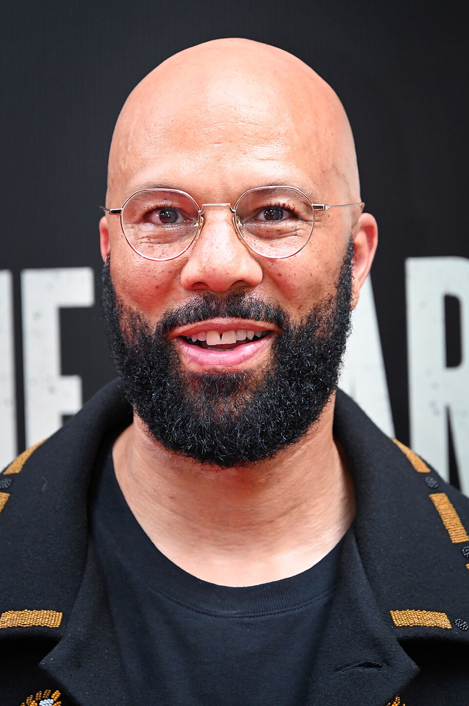 Close-up archival portrait of Common wearing round glasses at a Broadway opening night in April 2026.