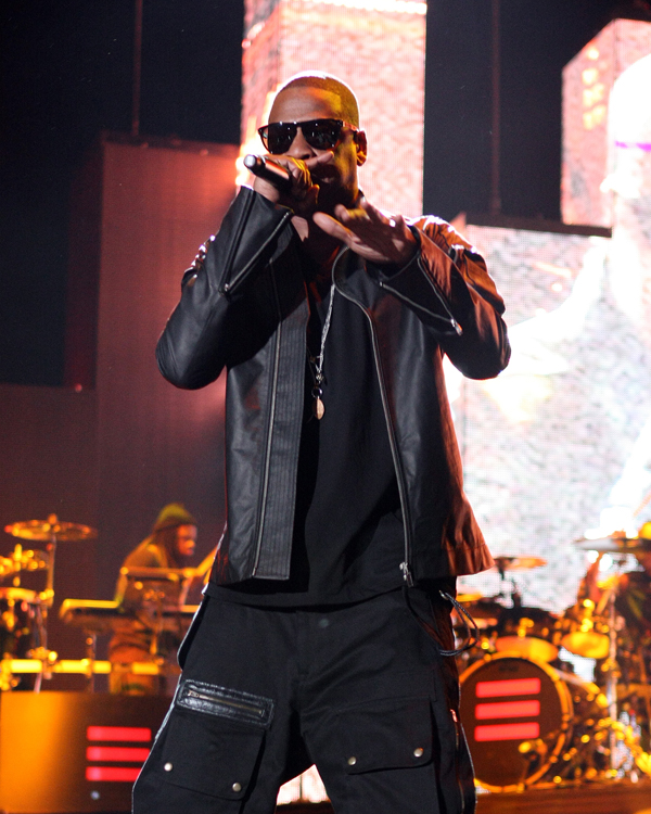 Jay-Z performs into a handheld microphone onstage, wearing black sunglasses and a black leather jacket, with band equipment and bright screens behind him.