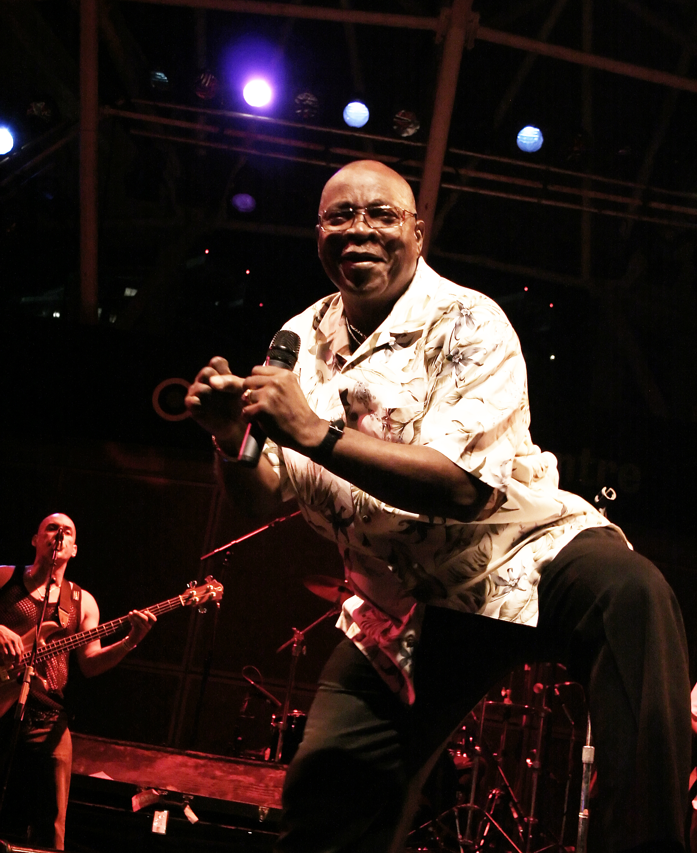 Mighty Sparrow leans toward the audience while singing into a handheld microphone onstage.