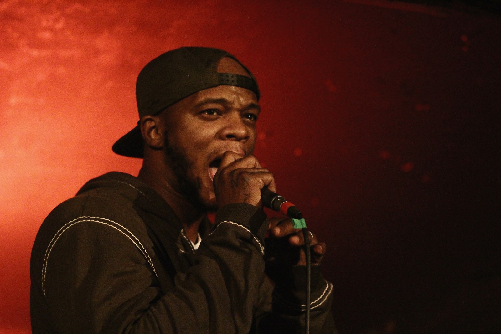 Papoose performing with a handheld microphone, wearing a backward black cap.